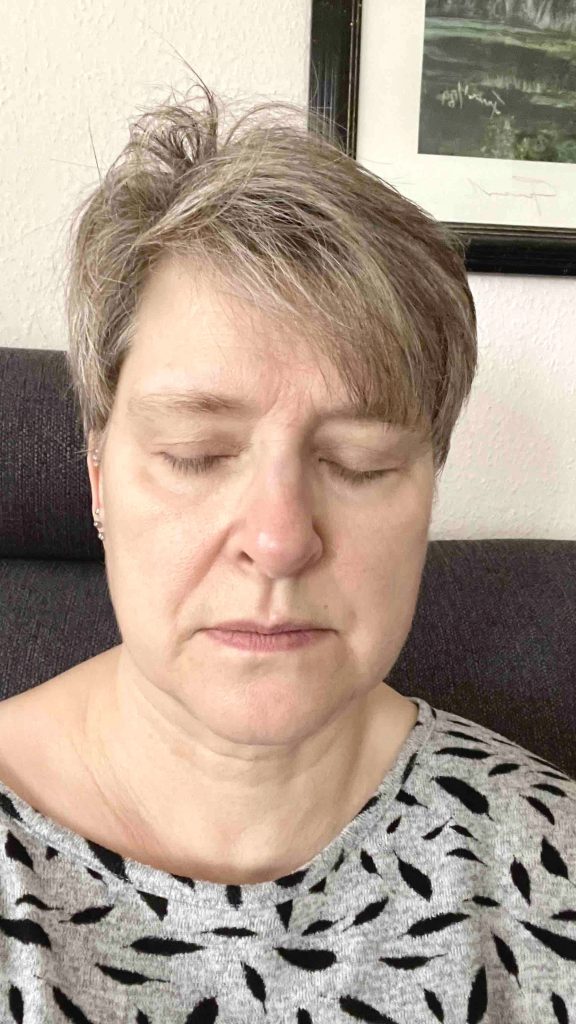 Frau mit grauem Shirt und geschlossenen Augen sitzt auf einem Sofa und meditiert anscheinend
